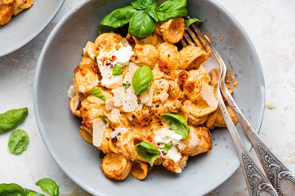 A bowl of pasta topped with creamy sauce, fresh basil leaves, and grated cheese. Two ornate forks rest on the side of the bowl. The background is light and scattered with additional basil leaves.