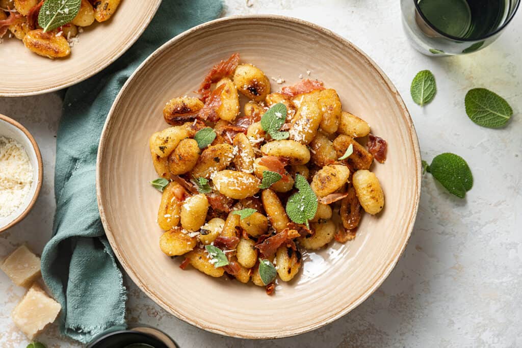 A bowl of crispy gnocchi garnished with fresh sage leaves, grated Parmesan, and crispy prosciutto. The bowl is set on a light surface, accompanied by a green cloth napkin. Additional bowls of gnocchi, Parmesan, and sprigs of sage are in the background.