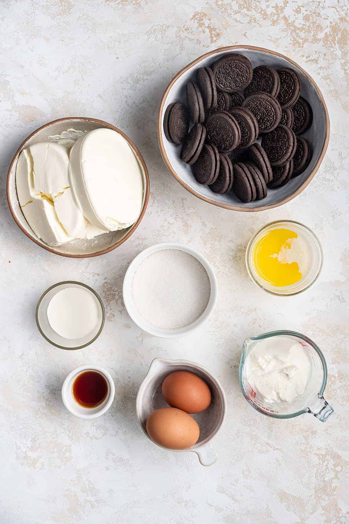 A bowl of Oreos, a bowl of each cream cheese, sour cream, heavy cream, sugar, melted butter, two eggs and vanilla extract on a white counter top.