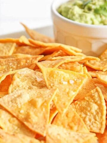 A pile of gluten-free homemade corn tortilla chips on a white platter with a side of guacamole in a small white bowl.