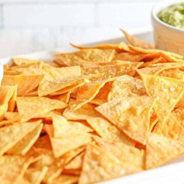A pile of gluten-free homemade tortilla chips on a white platter with a side of salsa and guacamole.