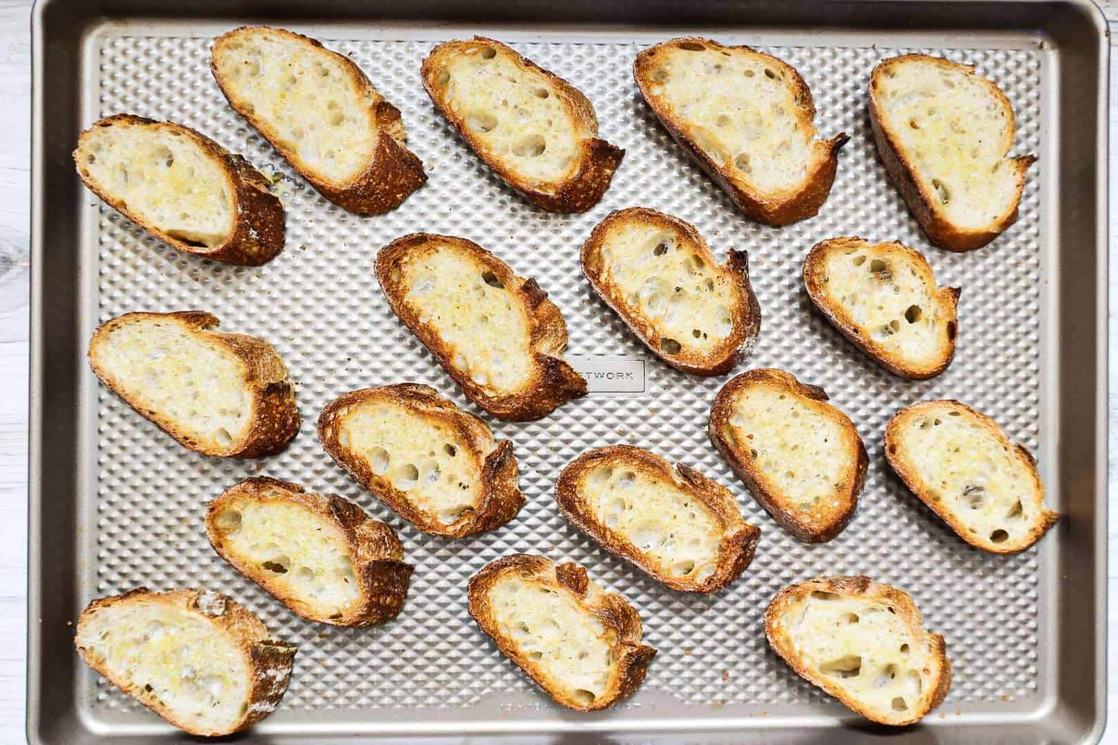 Seventeen pieces of toasted crostini on a large baking sheet.