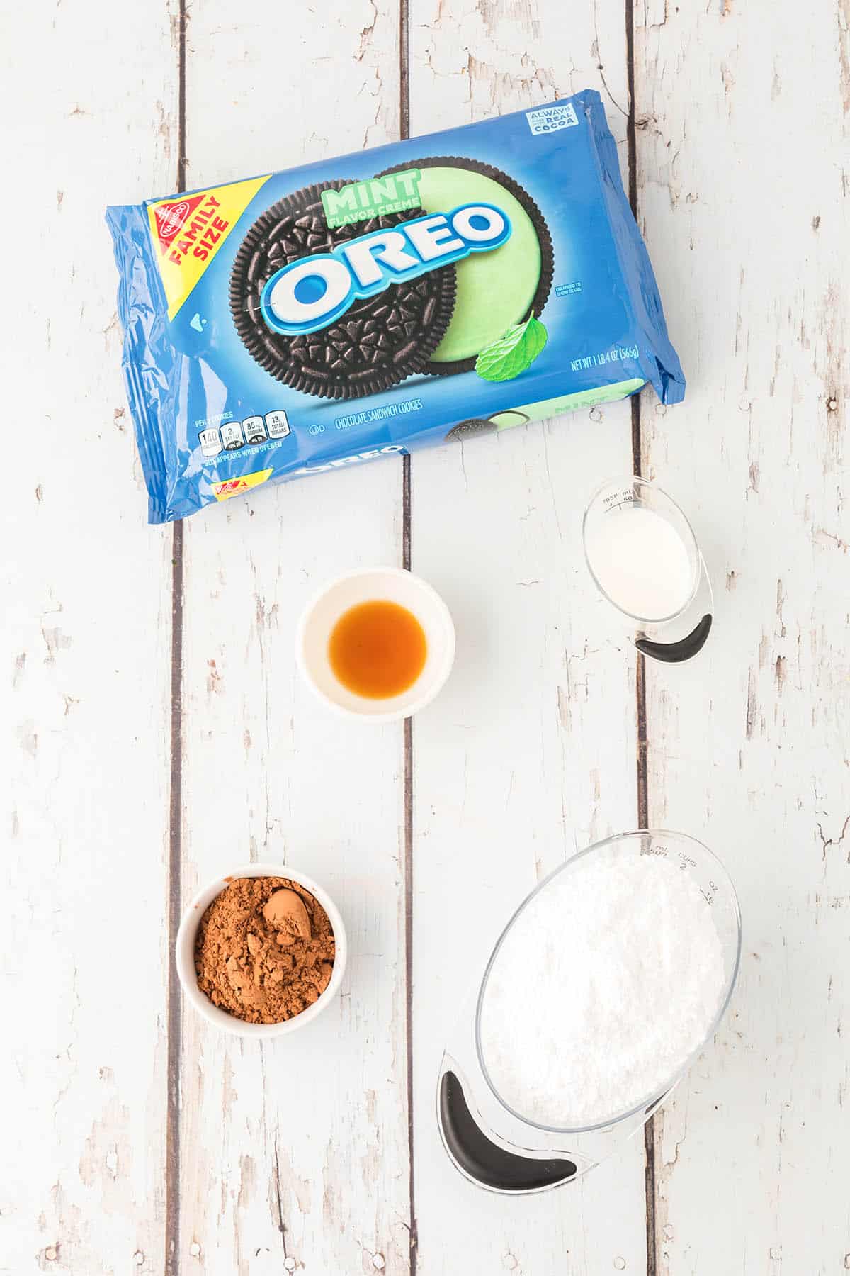 A package of mint oreo cookies, a measuring cup of powdered sugar, a ramekin of cocoa powder, a small bowl of vanilla extract and a small measuring cup of milk on a wooden countertop.