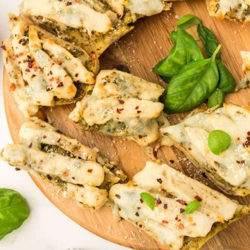 Several pieces of chicken pesto flatbread pizza garnished with fresh basil and red pepper flakes on a round wooden cutting board on a white counter top.