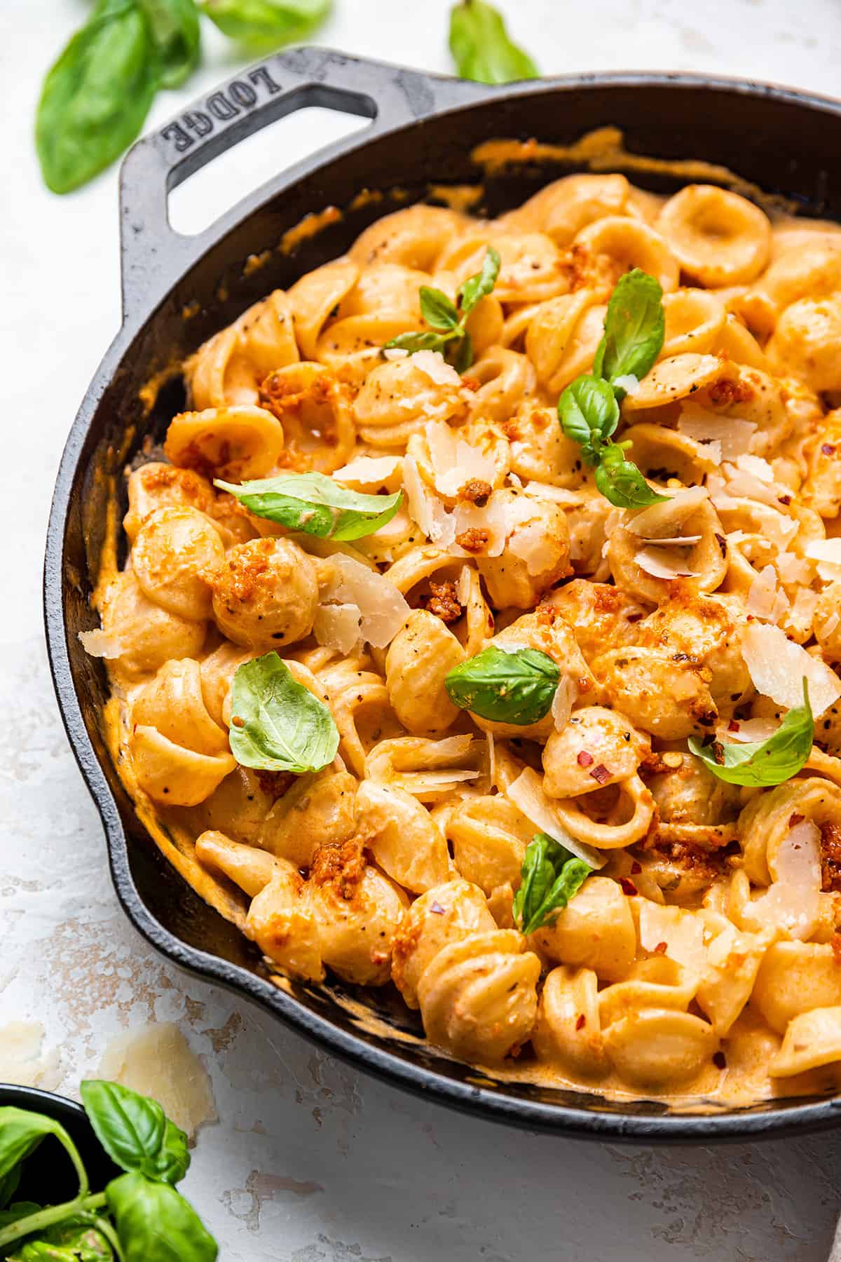 A large pan with a creamy sun dried tomato pesto sauce mixed with orecchiette pasta garnished with fresh basil leaves on a white counter top.