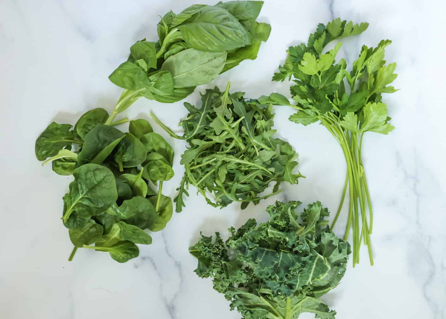 A handful of each fresh basil, spinach, arugula, kale, and parsley on a white countertop.