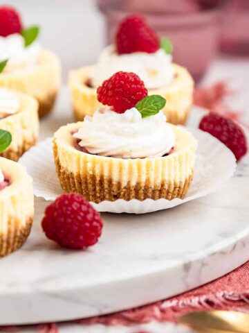 Five mini raspberry cheesecakes topped with whipped cream, a fresh raspberry and a mint leave on a white plate.