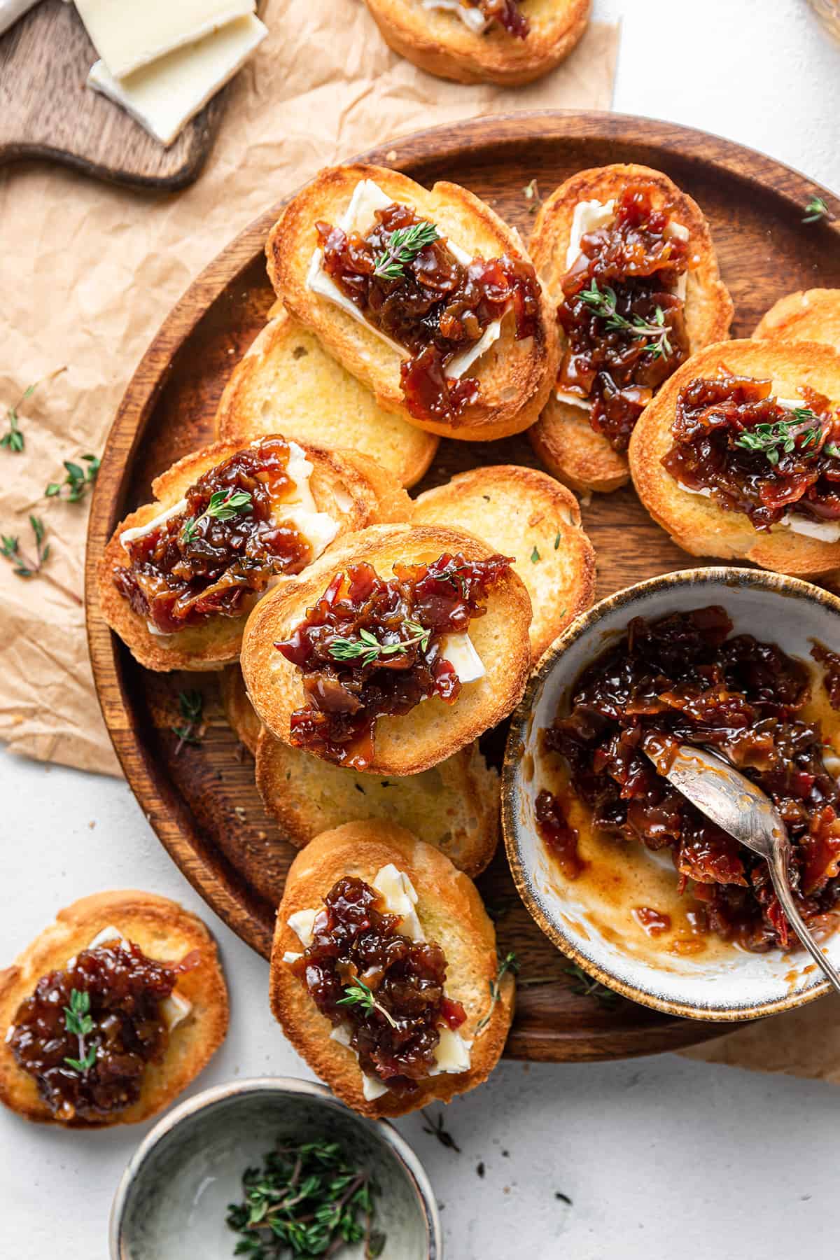 Several pieces of bourbon bacon jam crostini topped with fresh thyme on a circular wooden serving board next to a bowl of bacon jam.