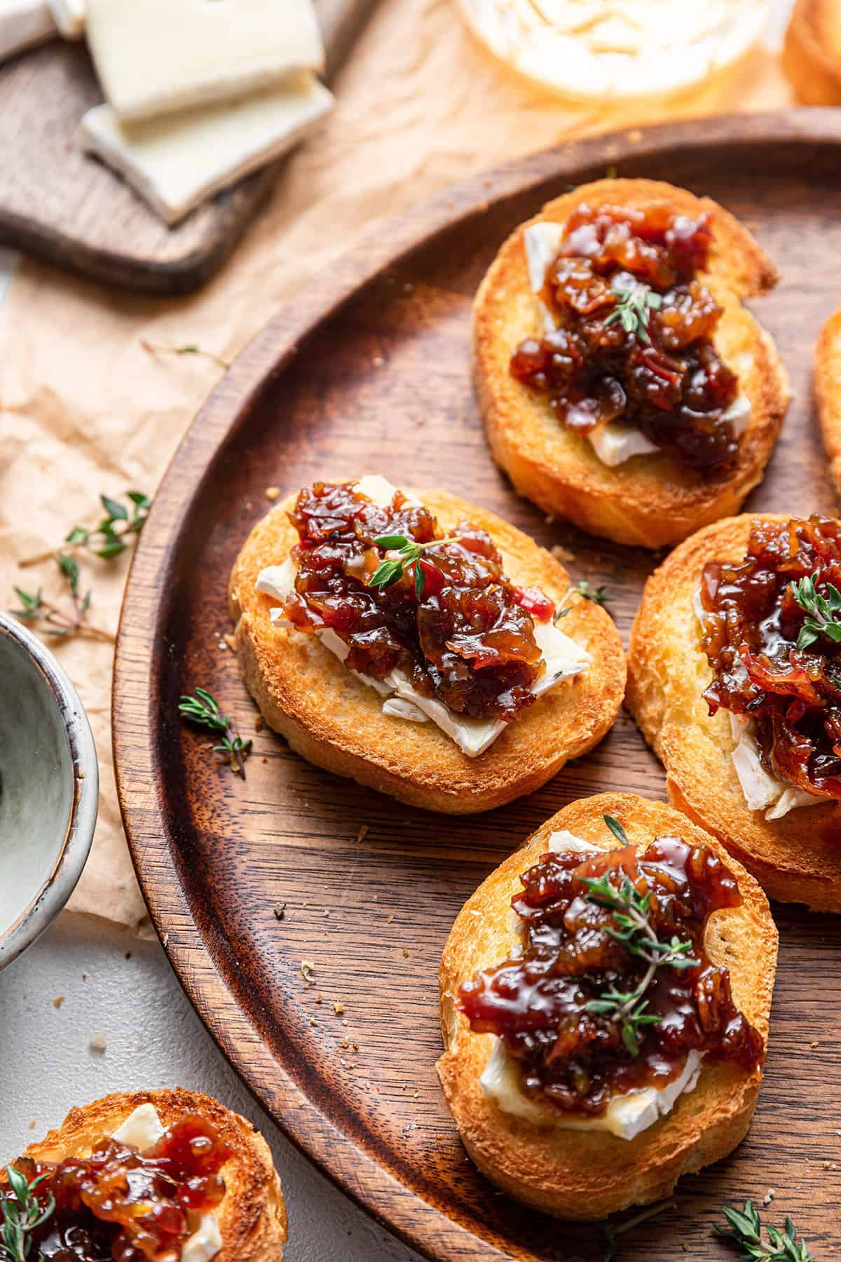 Four pieces of bourbon bacon jam crostini on a circular wooden plater and garnished with fresh thyme.