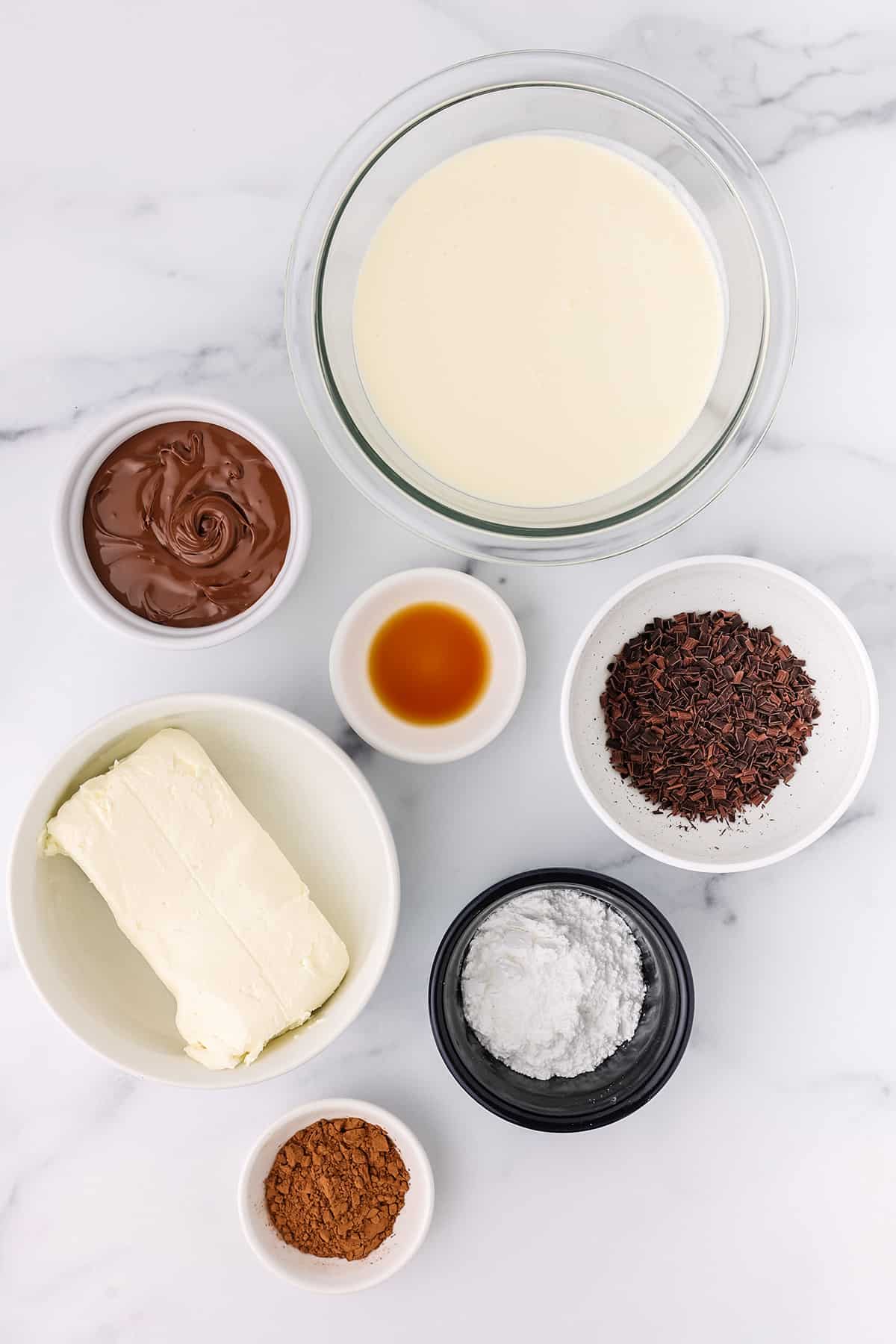 A bowl of heavy cream, cream cheese, cocoa powder, vanilla extract, Nutella, powdered sugar, and chocolate shavings on a white countertop.