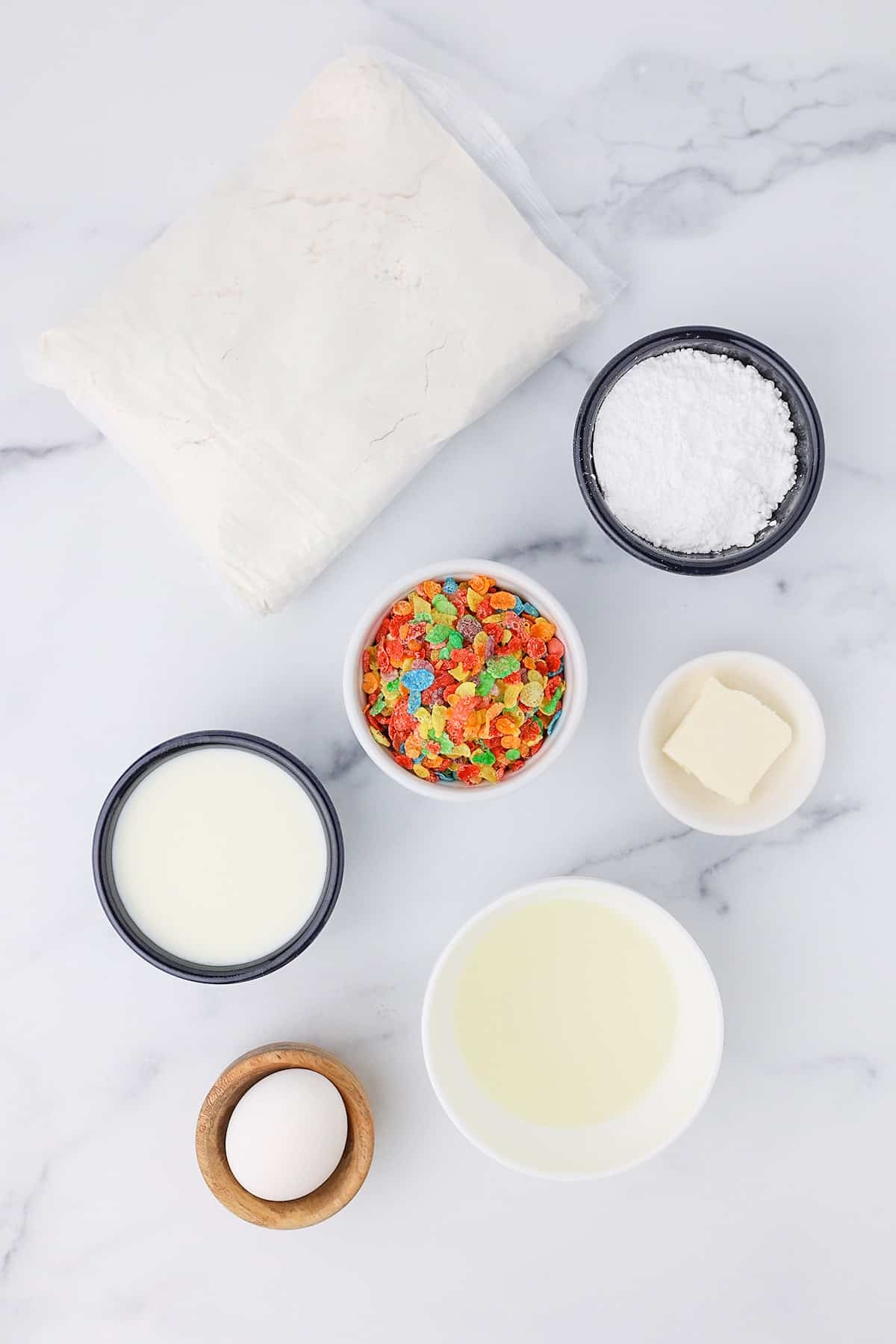 A package of white cake mix, some powdered sugar, a small bowl of fruity pebbles cereal, whole milk, one table spoon of butter, vegetable oil and a large white egg on a white counter top.
