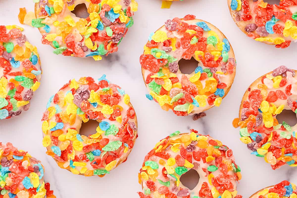 Nine frosted baked cake mix donuts topped with fruity pebbles on a wire rack on a white countertop.