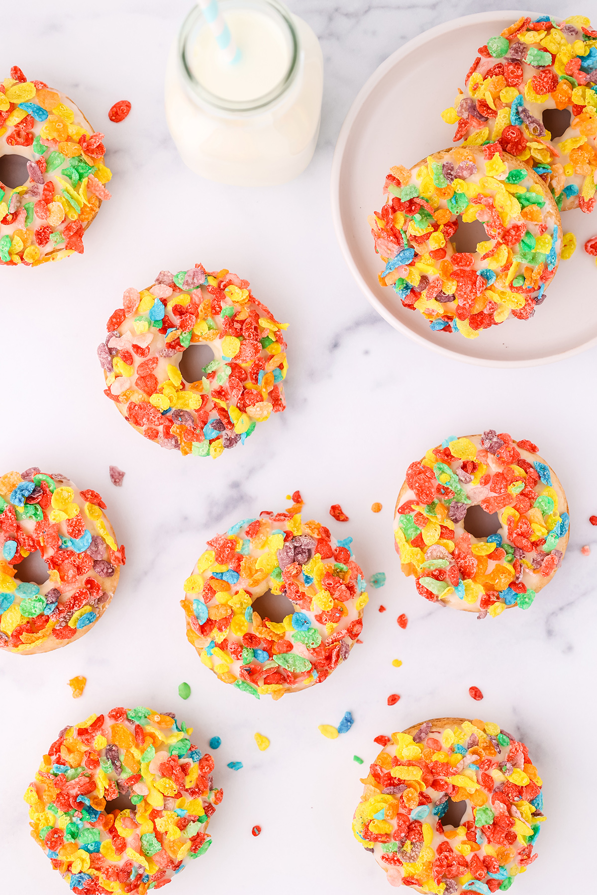 Seven fruity pebbles donuts on a white counter top and two on a white plate with a glass of milk on a white countertop.