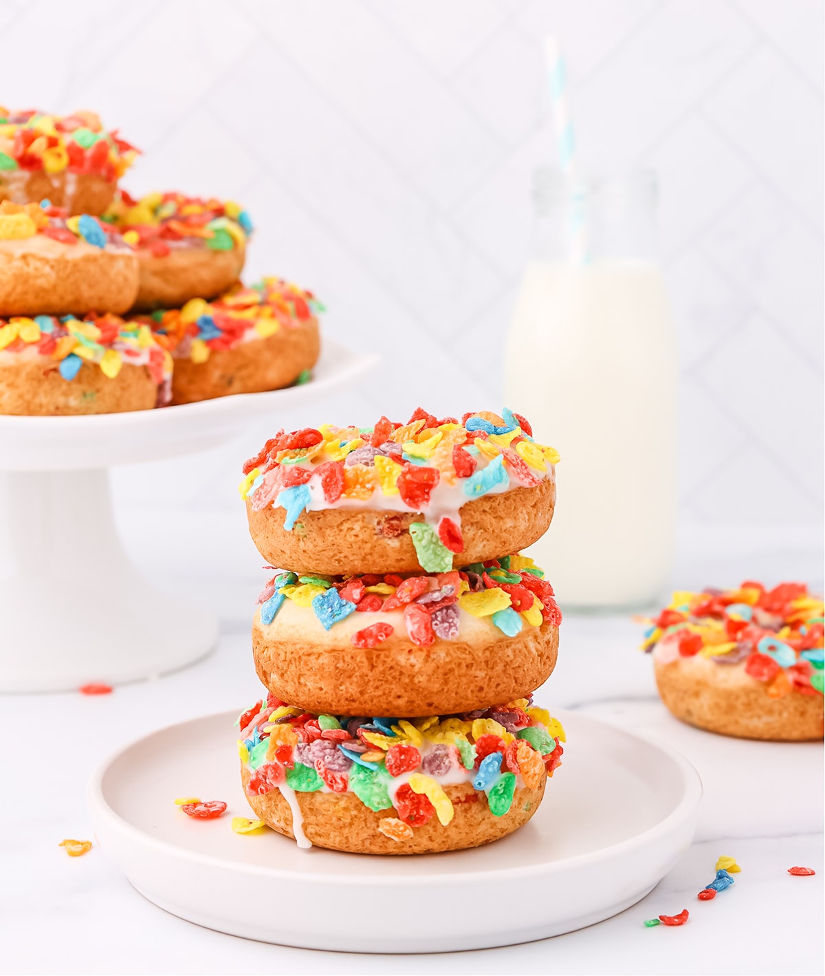 A stack of three fruity pebbles donuts on top of each other on a white plate with a cake stand holding more donuts and glass of milk in the background.