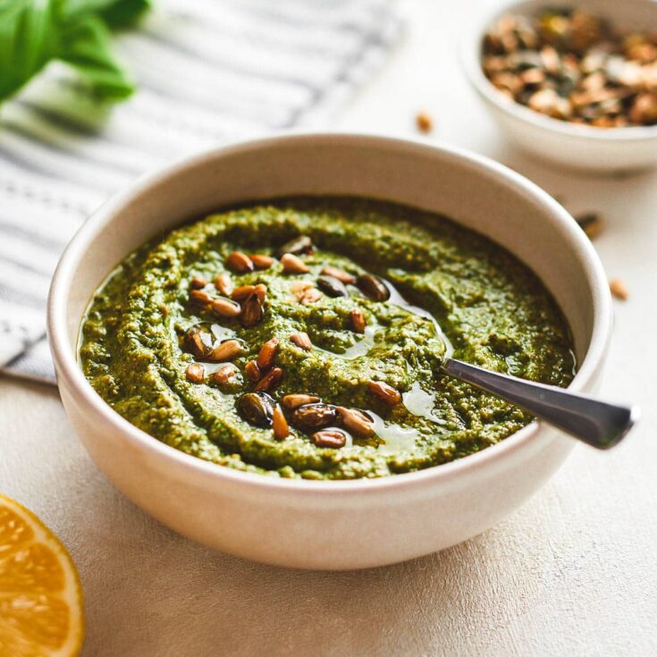 A large white bowl of zucchini pesto on a white counter top with a silver spoon in it.