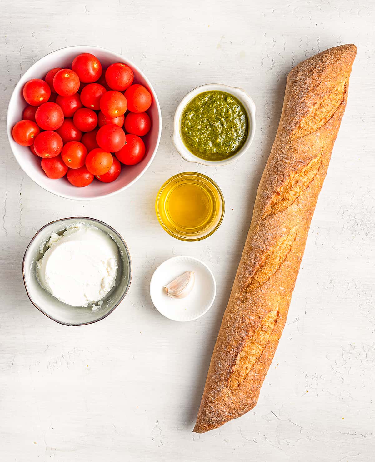 A bowl of cherry tomatoes, a long baguette, ricotta cheese, a garlic clove, pesto and olive oil on a white counter top.