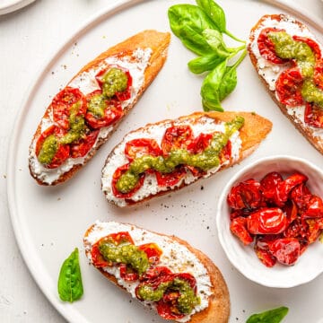 Four pieces of pesto ricotta crostini on a white plate with a small white bowl of slow roasted tomatoes on the same plate and fresh basil leaves scattered about.