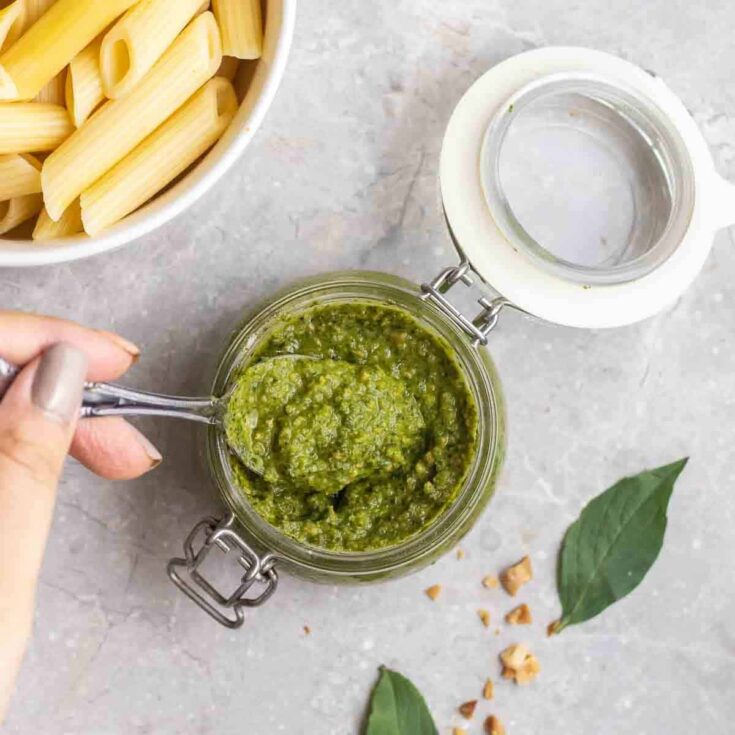 A small glass jar of thai basil pesto on a gray counter top being spooned out next to a bowl of penne pasta.