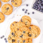 A large stack of lemon blueberry oatmeal cookies on a white cake stand with a white container of fresh blueberries and six more oatmeal cookies on a wire rack.