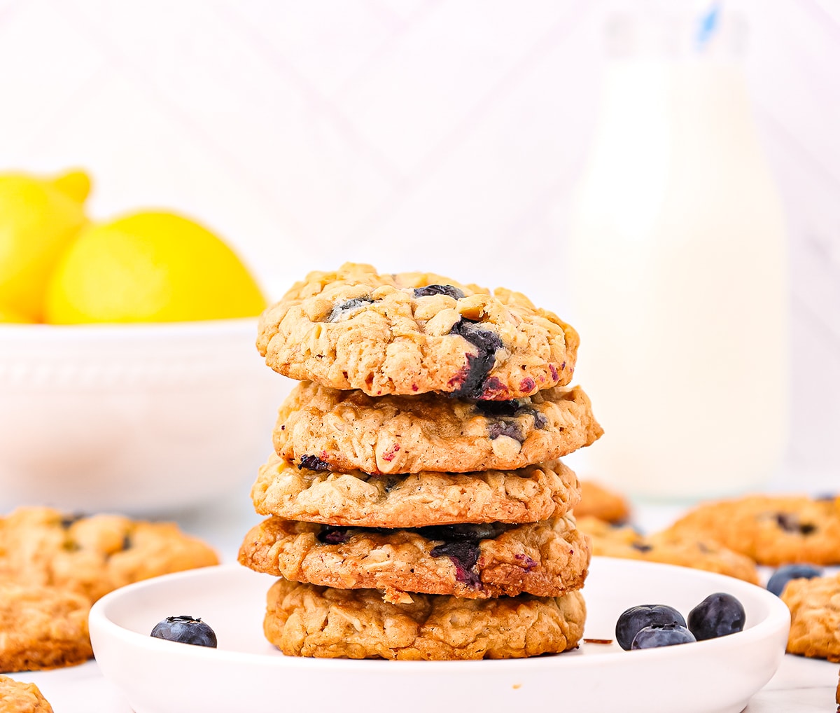 A stack of five blueberry lemon oatmeal cookies piled high on a white plate surrounded by fresh blueberries and a bowl of lemons and tall glass of milk in the background.