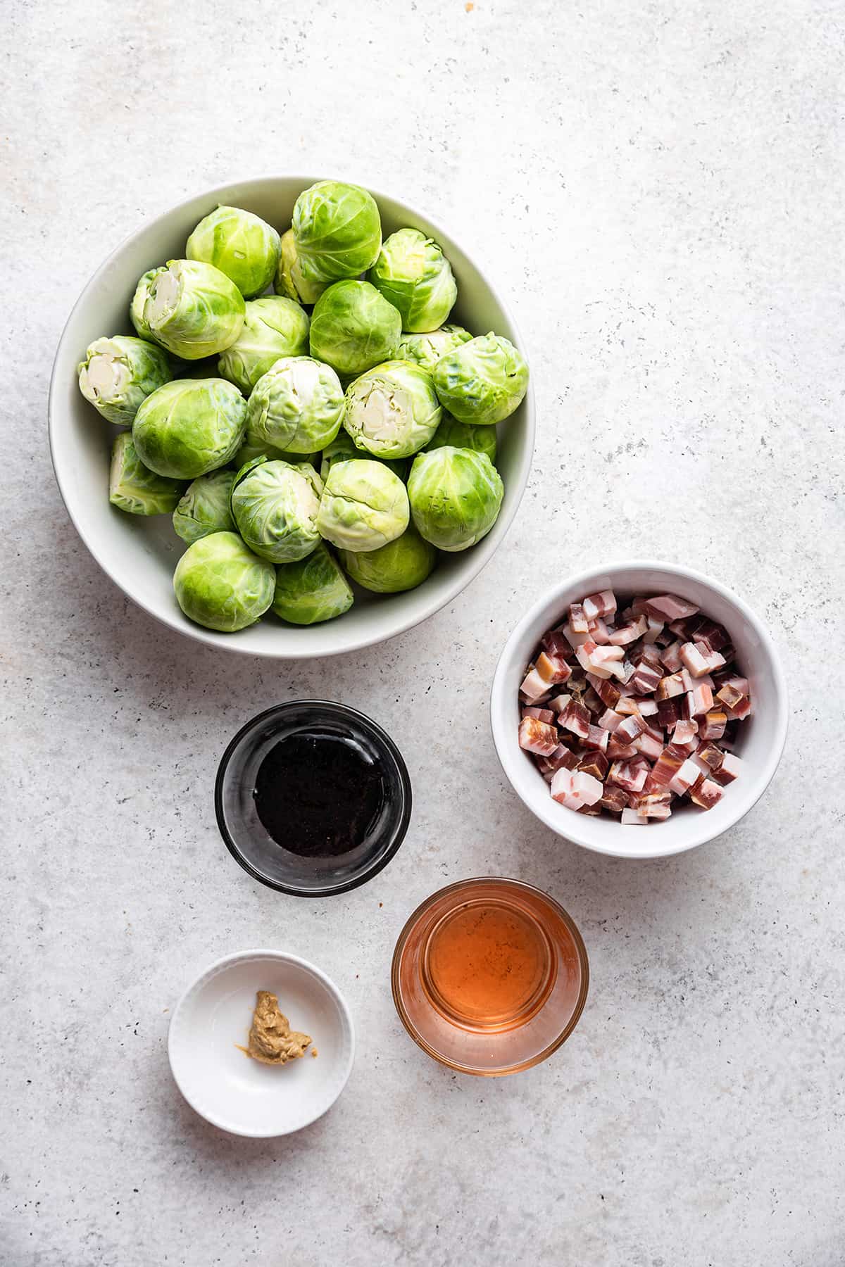 A bowl of fresh Brussels sprouts, balsamic glaze, red wine vinegar, dijon mustard and diced pancetta on a white countertop.