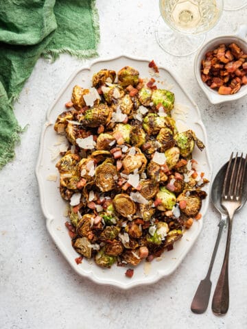 A large white plater with flash fried Brussels sprouts with crispy pancetta and parmesan on a white counter top with a green napkin nearby.