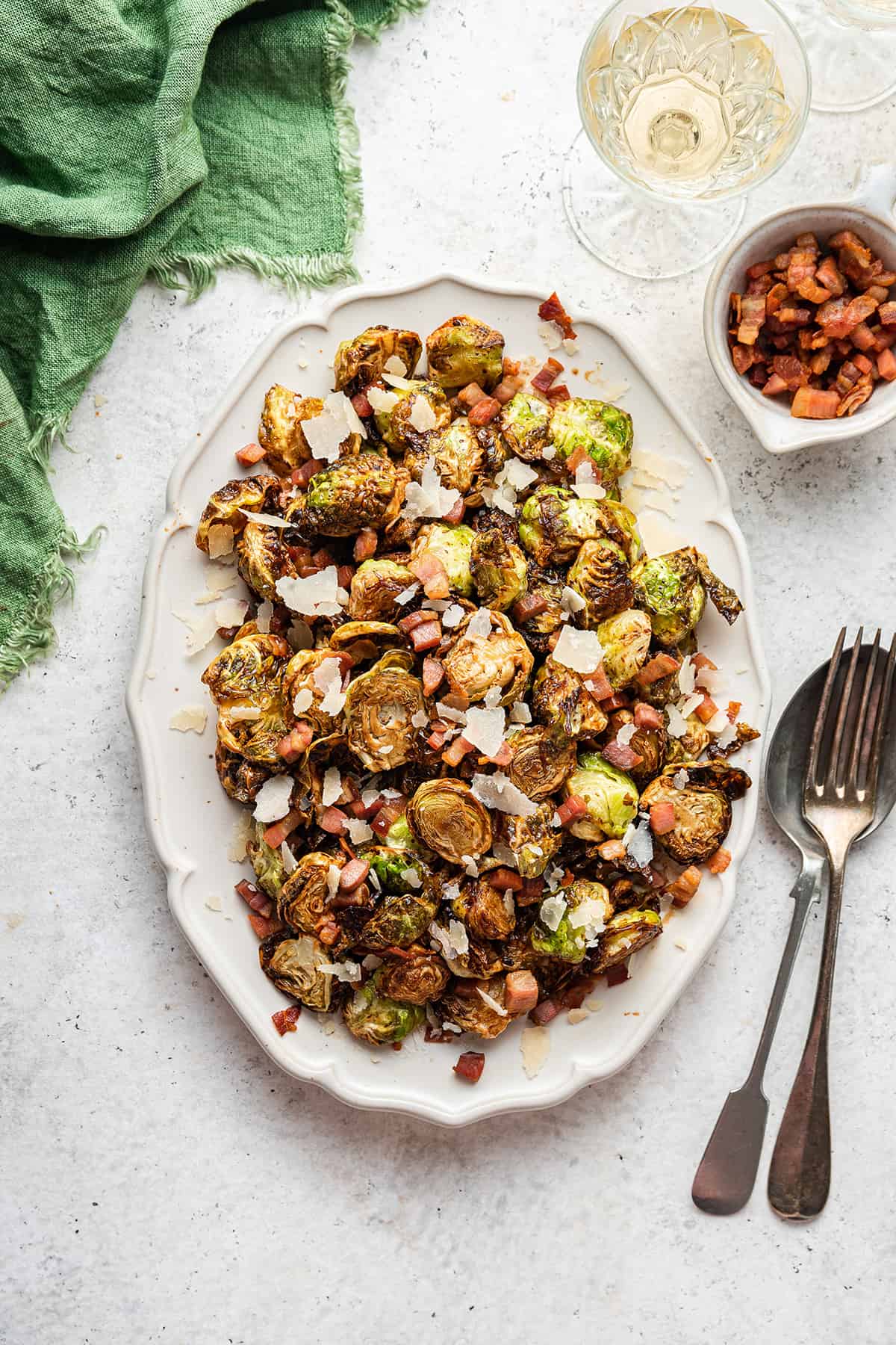 A large white plater with flash fried Brussels sprouts with crispy pancetta and parmesan on a white counter top with a green napkin nearby.