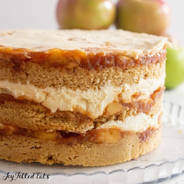 A large apple cider donut cake on a glass platter with whole green apples in the background.