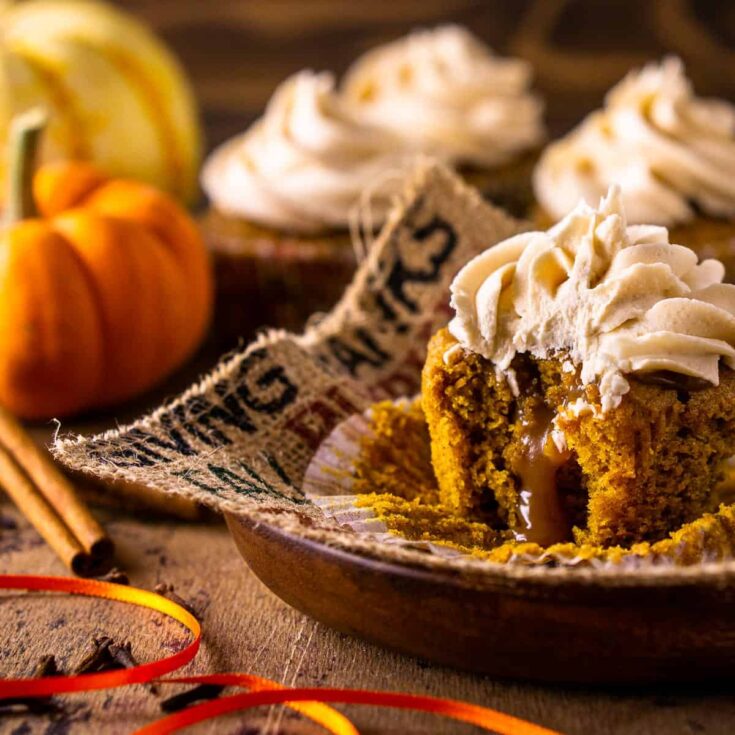 A brown butter pumpkin cupcake with a bite taken out of it on a brown serving platter with more cupcakes and a pumpkin in the background.