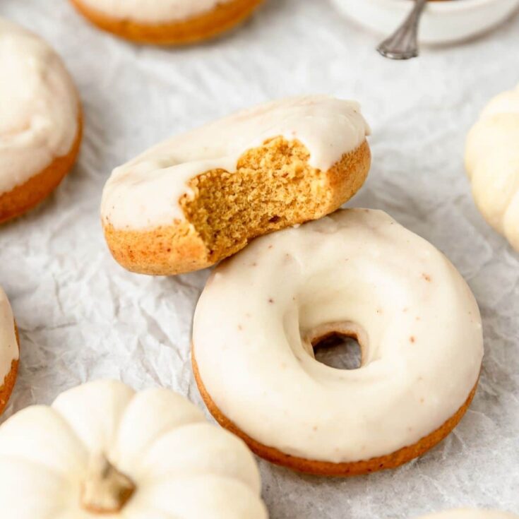 Two pumpkin donuts with cream cheese frosting leaning against each other with one missing a bite out of it on white parchment paper.