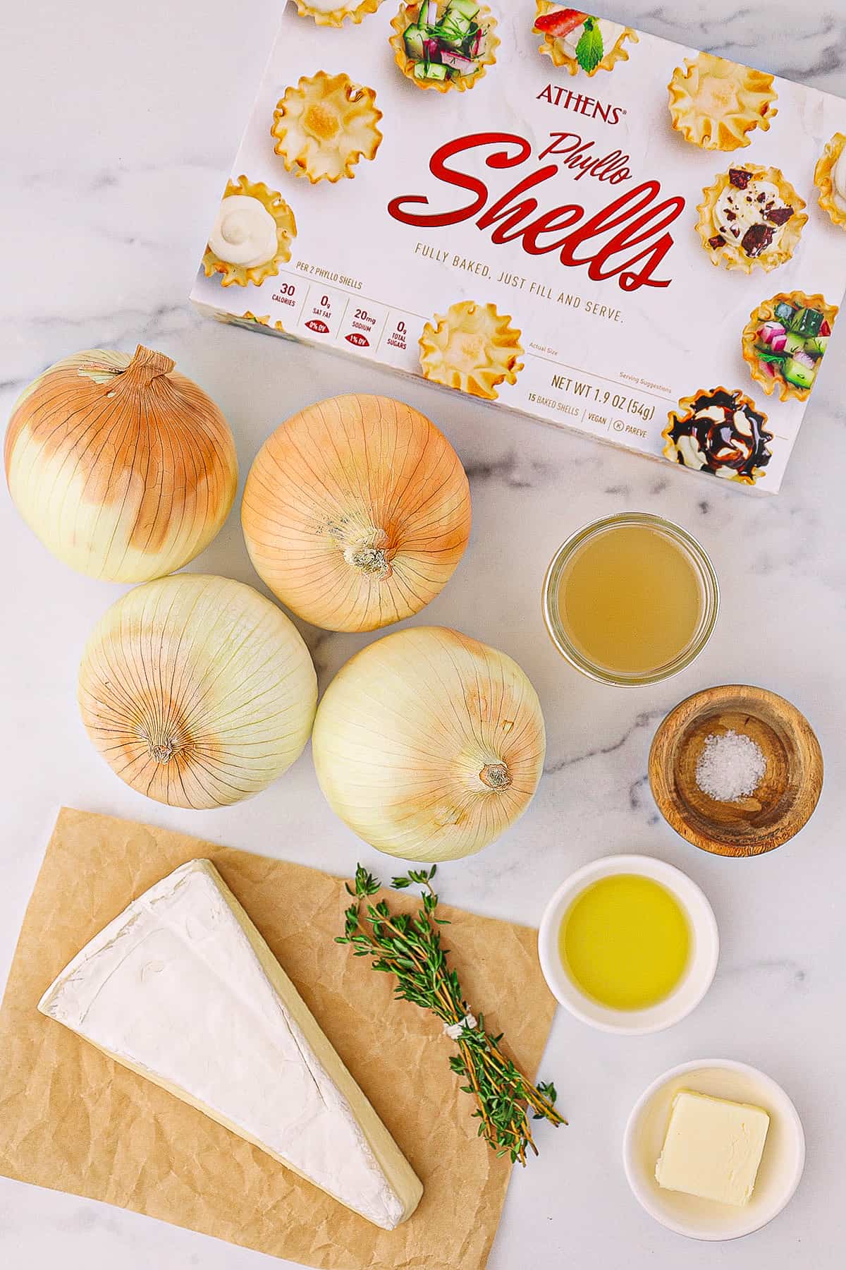 An unopened box of phyllo shells, four whole onions, a large wedge of brie cheese, a small glass container of chicken broth, salt, fresh thyme, olive oil , and butter on a white counter top.