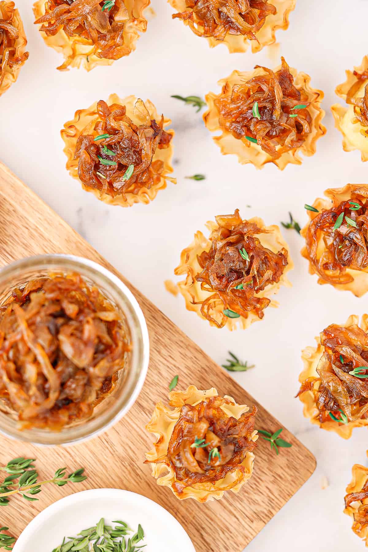 Eight caramelized onion tartlets on a white counter top with a jar of caramelized onions and a white ramekin of fresh thyme on a wooden cutting board.