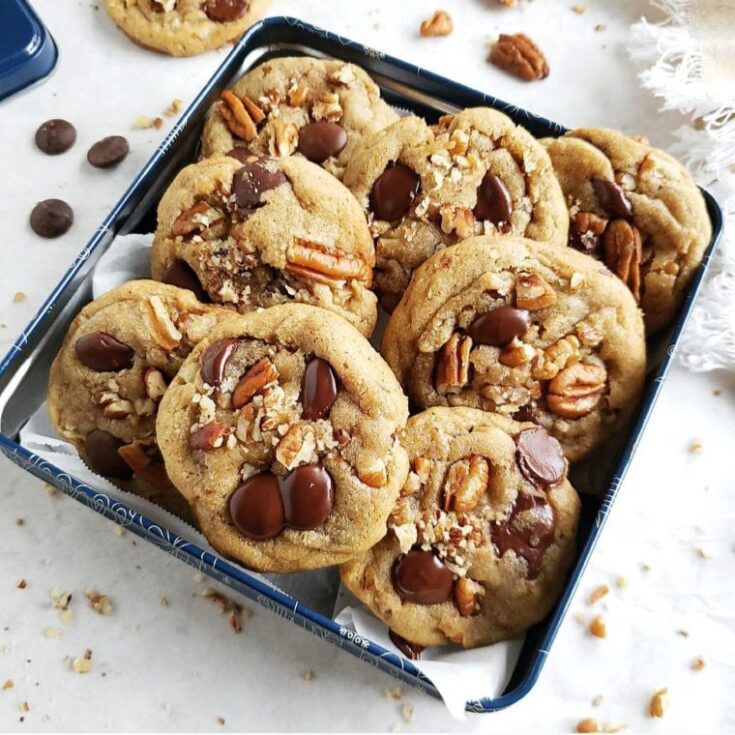 Eight chocolate chip pecan cookies in a blue cookie tin on a white counter top.
