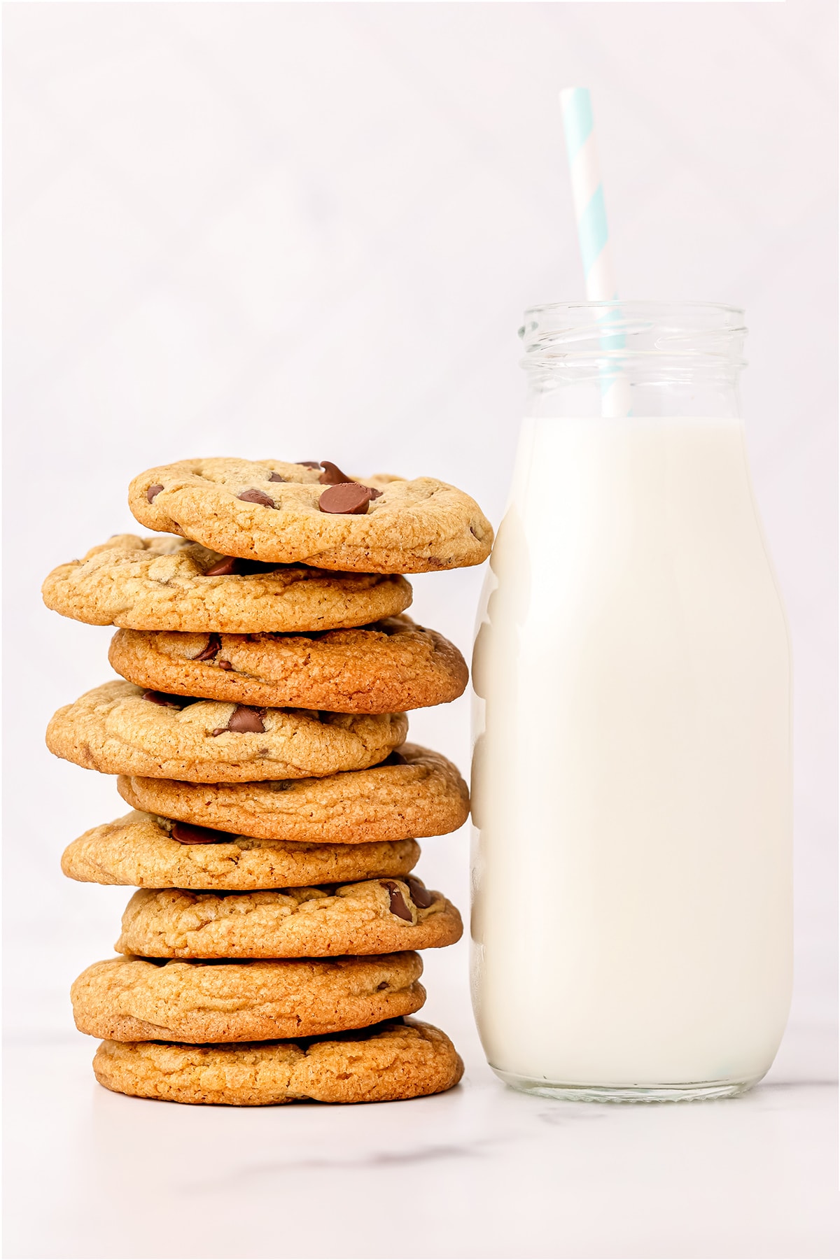 A large tower of chocolate chip cookies stacked on top of each other leaning next to a tall glass of milk with a blue striped straw.