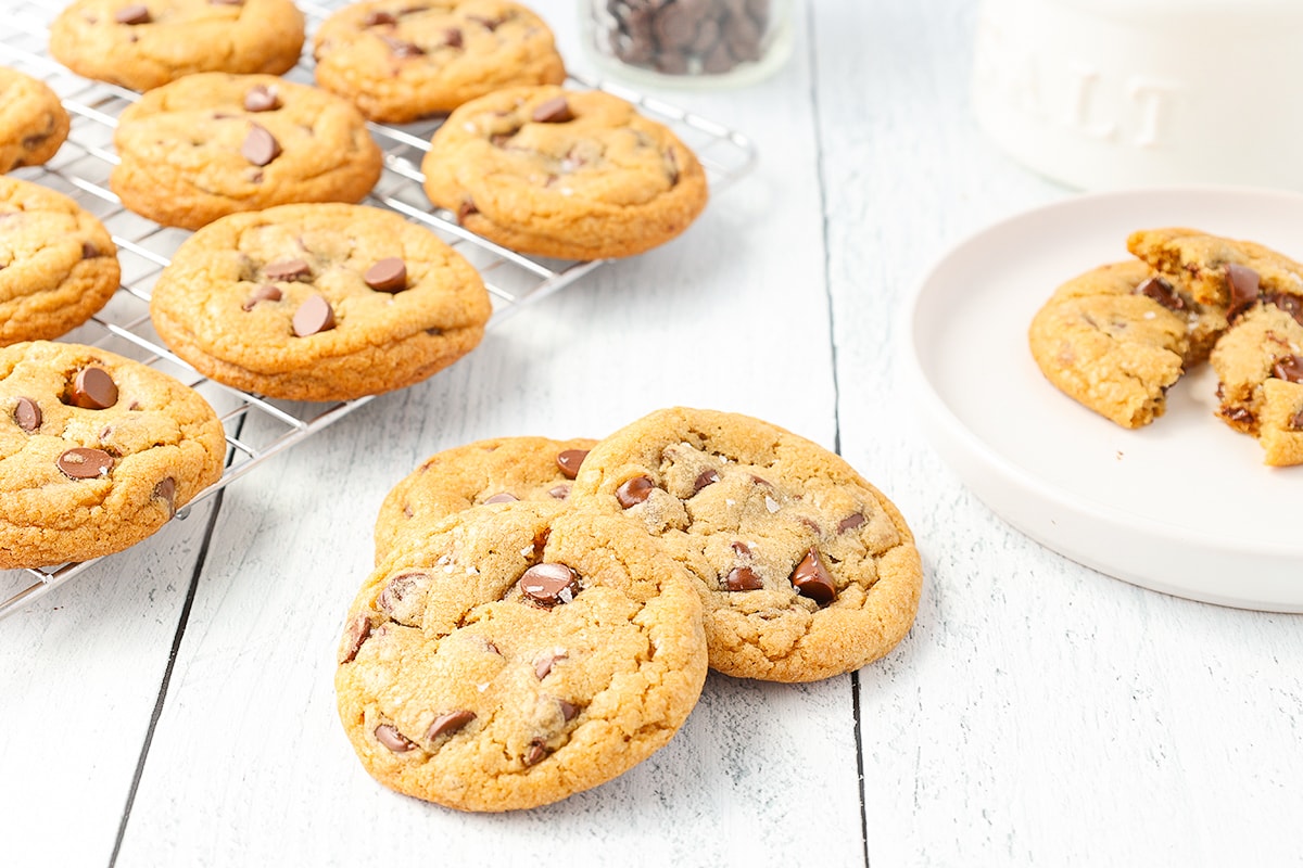 Several chocolate chip cookies resting on a wire rack and three cookies leaning on each other with one cookie broken on a white plate.