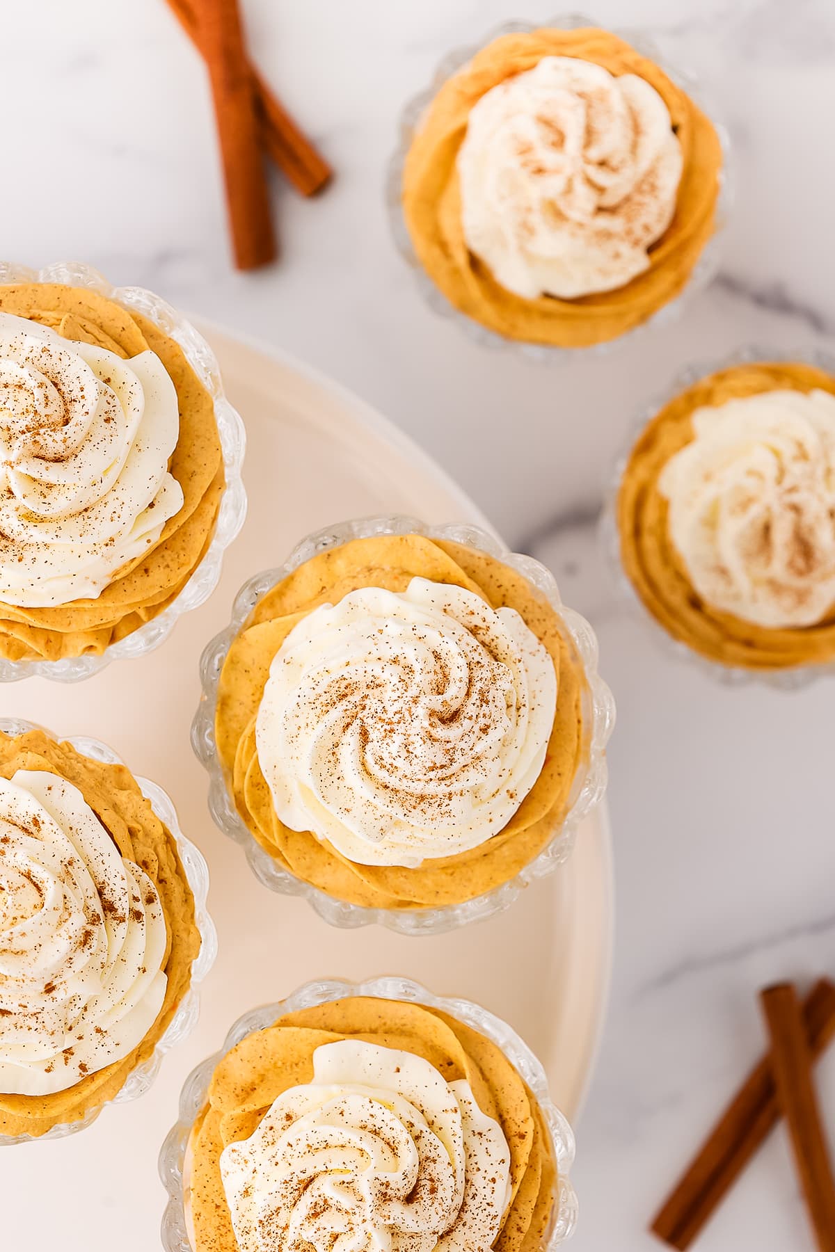 Four dessert cups filled with pumpkin cheesecake mousse on a white cake stand with two cups of pumpkin mousse on the counter with whole cinnamon sticks scattered about.