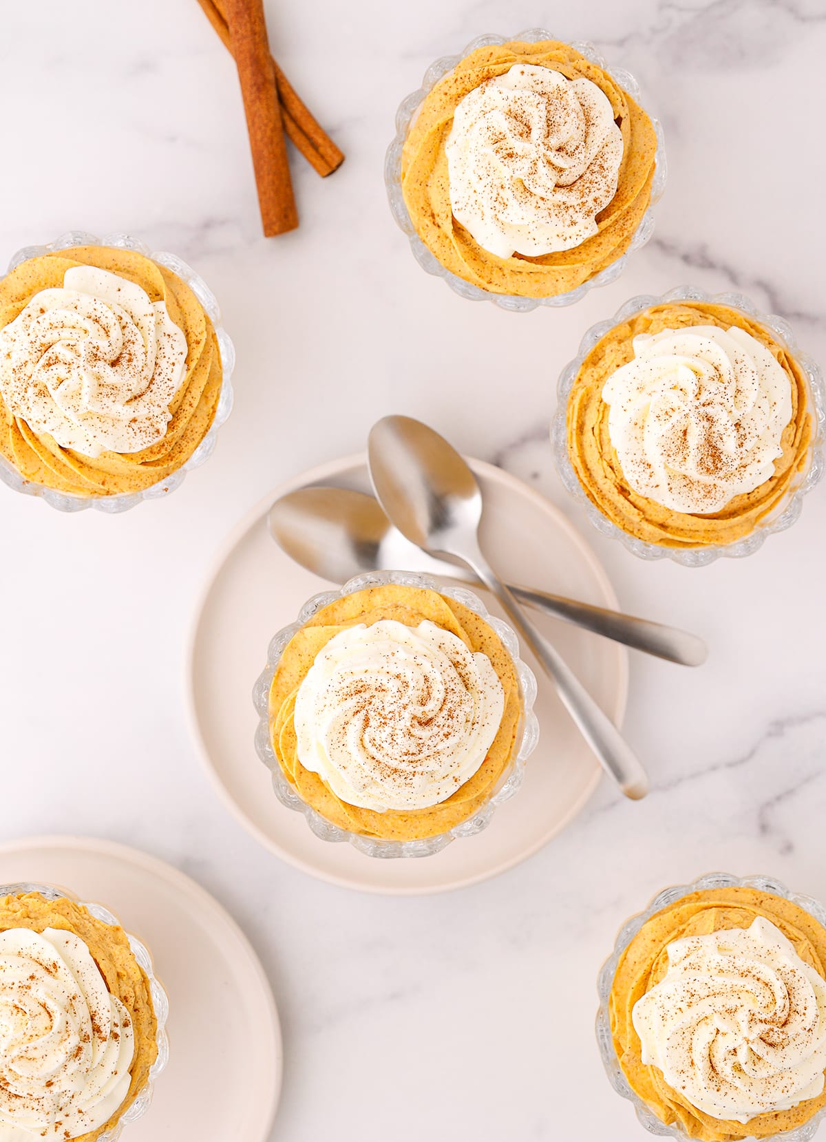 Four dessert cups filled with pumpkin mousse on a white counter and two more on white plates with two silver spoons laying nearby.