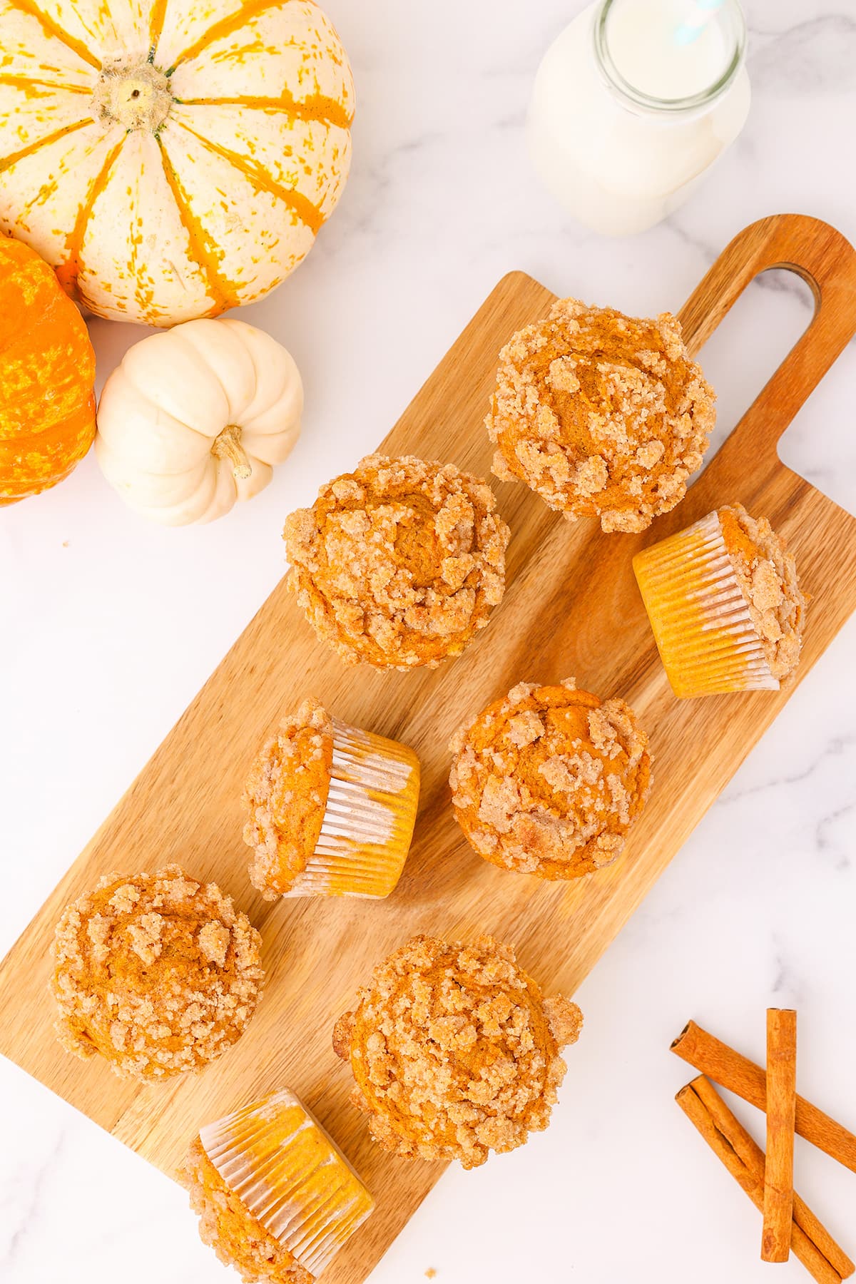 Eight pumpkin muffins on a long wooden cutting board with a few cinnamon sticks and three pumpkins laying nearby on a white counter top.