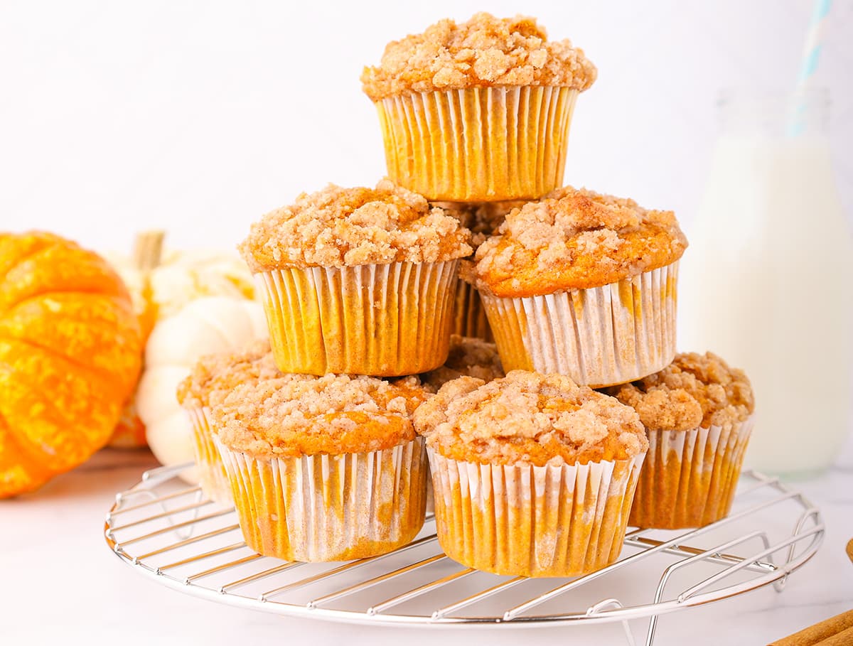 A tower of cake mix pumpkin muffins piled on top of a round metal cooling rack with three pumpkins and a tall glass of milk in the background.