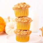 Three pumpkin muffins stacked on top of each other on a white plate with three pumpkins and a tall glass of milk in the background on a white counter top.