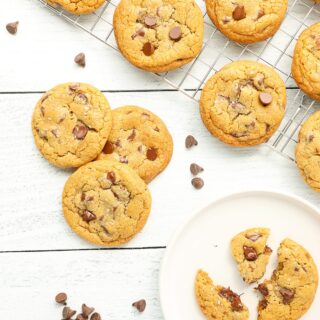 Several brown butter chocolate chip cookies cooling on a wire rack, three cookies leaning against each other on a white countertop and one cookie broken apart with melted chocolate chips on a white plate.
