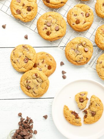 Several brown butter chocolate chip cookies cooling on a wire rack, three cookies leaning against each other on a white countertop and one cookie broken apart with melted chocolate chips on a white plate.