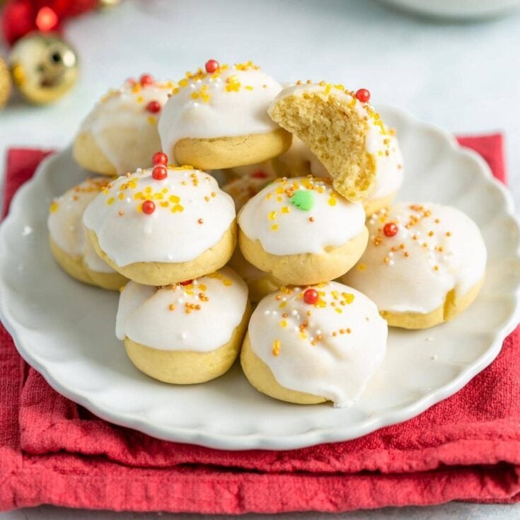 Several Italian anginetti Christmas cookies piled together on a white plate with a red napkin underneath on a white counter.