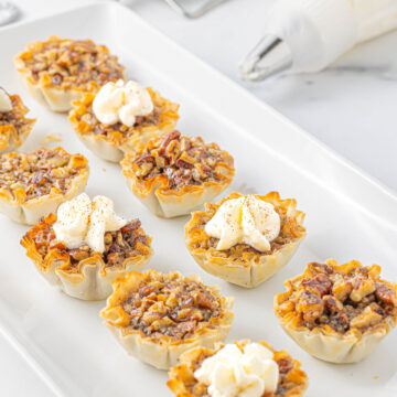 Several mini pecan pie bites on a white platter with a few topped with whipped cream and a piping bag of whipped cream resting next to the serving platter.