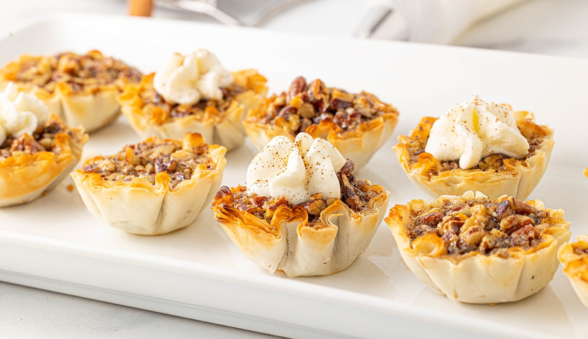 Eight pecan pie bites on a white rectangular serving platter with four topped with fresh whipped cream and cinnamon.