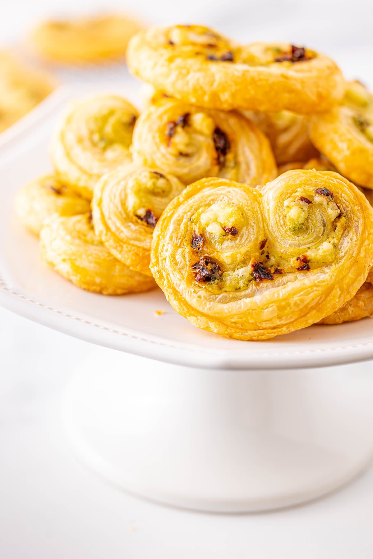A pile of sun dried tomato, pesto, and feta puff pastry palmiers on a white cake stand.