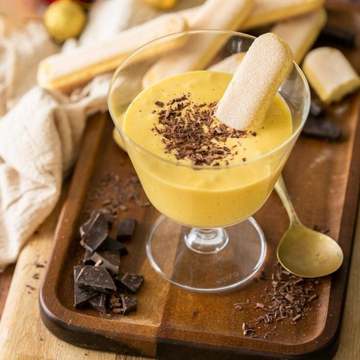 A small dessert glass filled with zabaglione on a wooden serving tray with ladyfinger cookies nearby.