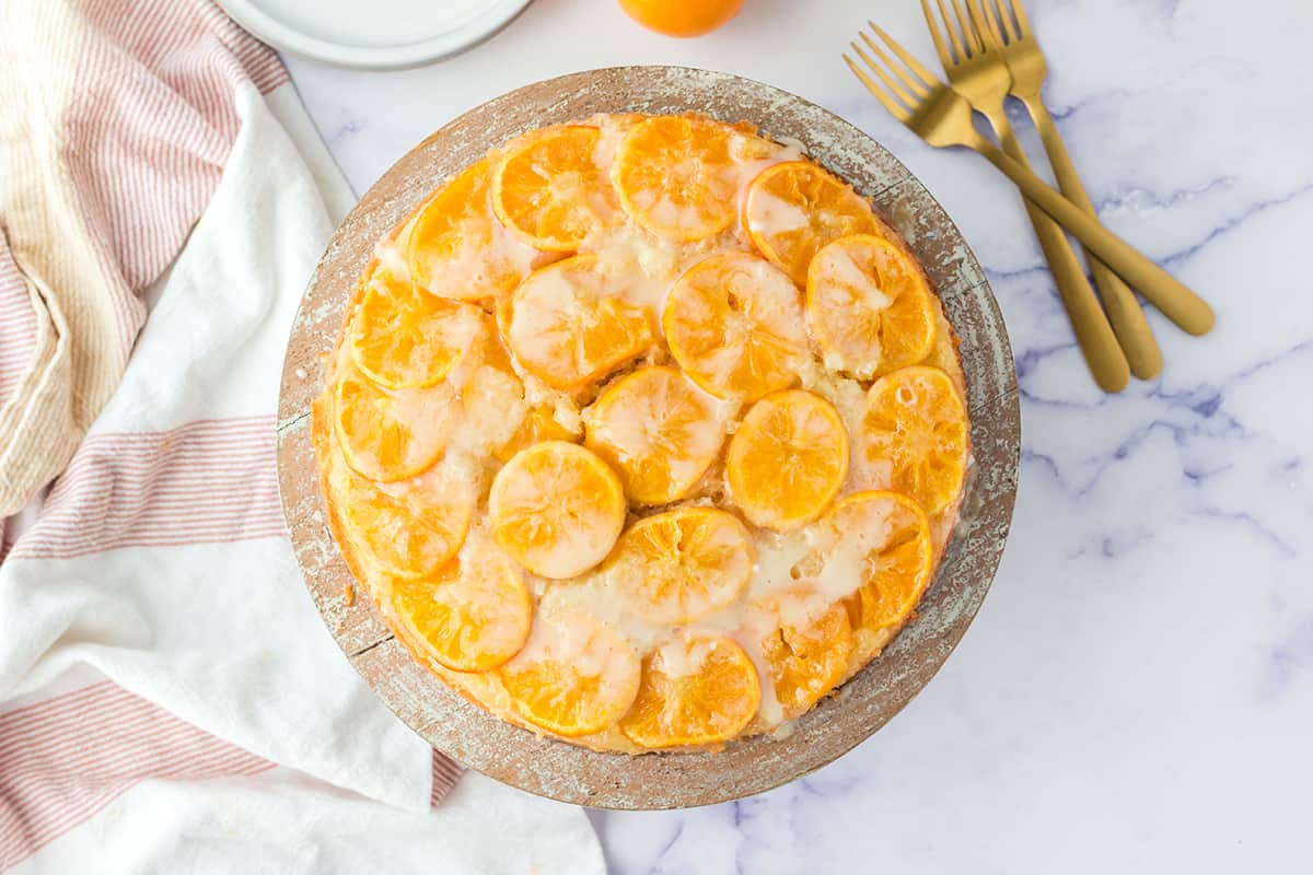 An orange upside down cake on a wooden cake stand with three gold forks nearby on a white counter top.