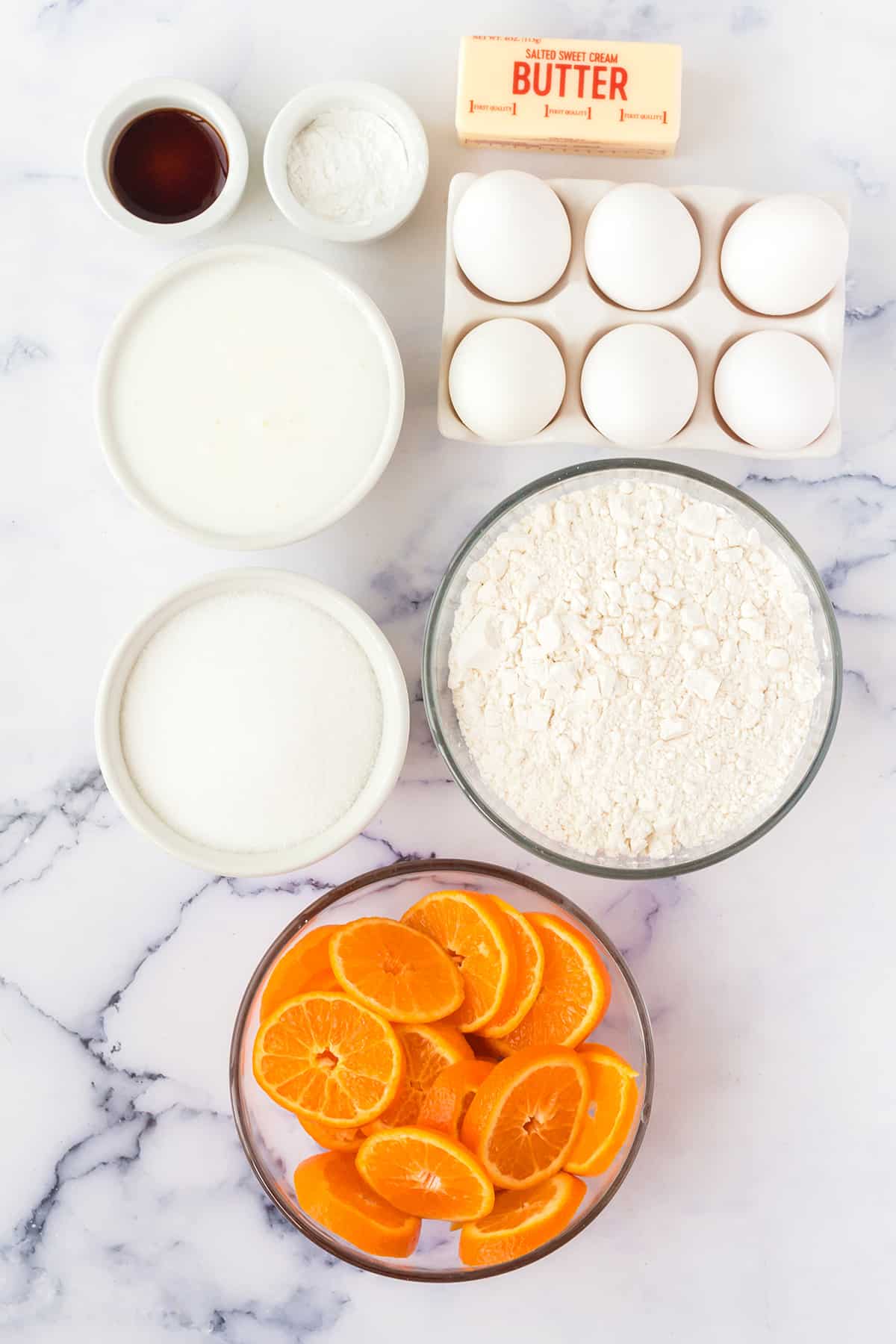 A bowl of sliced oranges, flour, granulated sugar, milk, eggs, baking powder and vanilla extract on a white counter top.