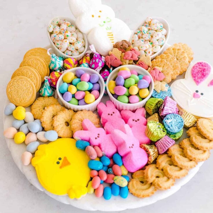 A white serving tray filled with marshmallow peeps, pastel colored candies, egg shaped chocolates, and bunny shaped crackers on a white counter top.